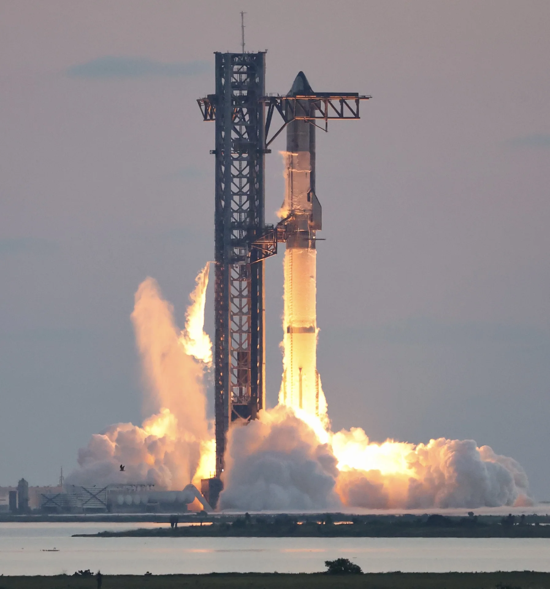 Starship stands beside its launch tower as orange engine flame and white exhaust spread across the pad.